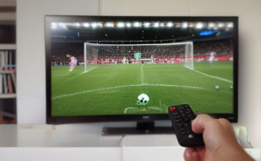 First person view of a man, his hand shown holding a remote while pointing at a TV. The TV display shows an ongoing soccer game.
